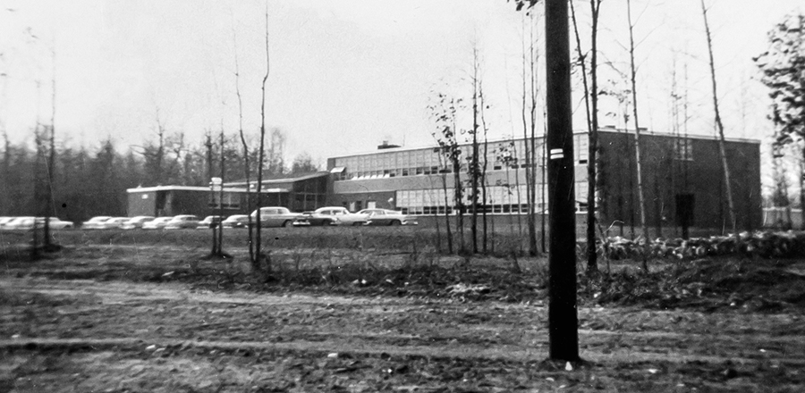 Black and white photograph of North Springfield Elementary School taken in 1958 for a fire insurance survey for the Fairfax County School Board. The school is seen from Heming Avenue and because there are so few trees at this time the rear of the building is visible. A two-story classroom wing and the cafeteria are visible from this angle. There is a row of 1950s era cars parked outside.
