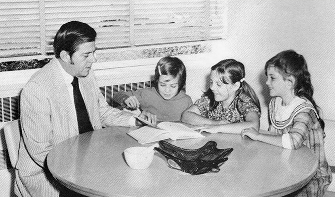 Black and white yearbook photograph of Principal Ronald E. West. He is seated at a table looking at a book with three students. 