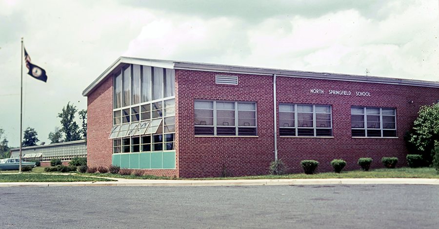 Color photograph from a 35 millimeter slide of the front of North Springfield Elementary School. A blue car is parked in front of the building. The windows are open because in these days the school did not have air conditioning. 