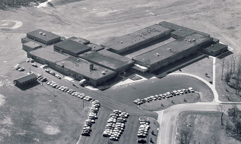 Black and white aerial photograph of Poe Intermediate School. It is an L-shaped building with several classrooms wings of both one and two-stories. The photograph was probably taken in the 1960s because the trees planted in front of the school are very small. 